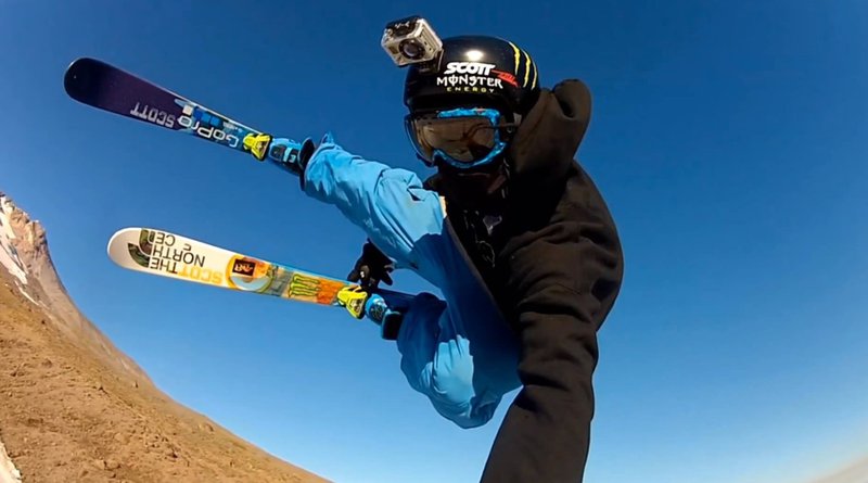 Person skiing with a GoPro camera mounted on their helmet, performing a mid-air trick against a clear blue sky.